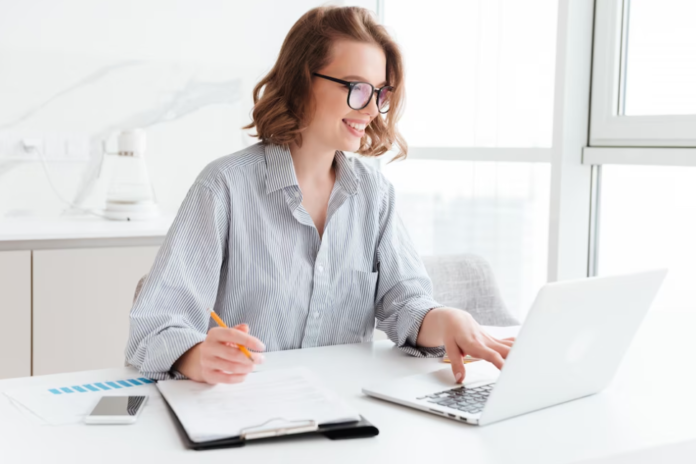 woman in shirt and glasses, holding a pen in one hand and typing with another hand