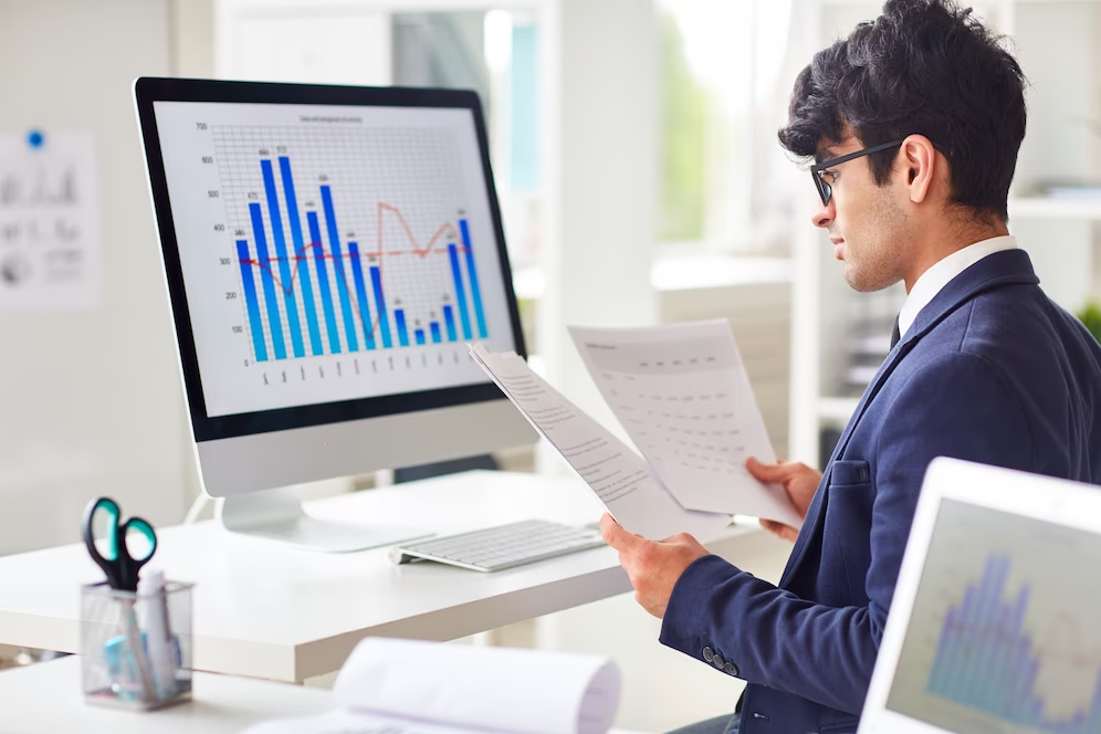 Man sitting in front of a computer, displaying graphs, and holding documents